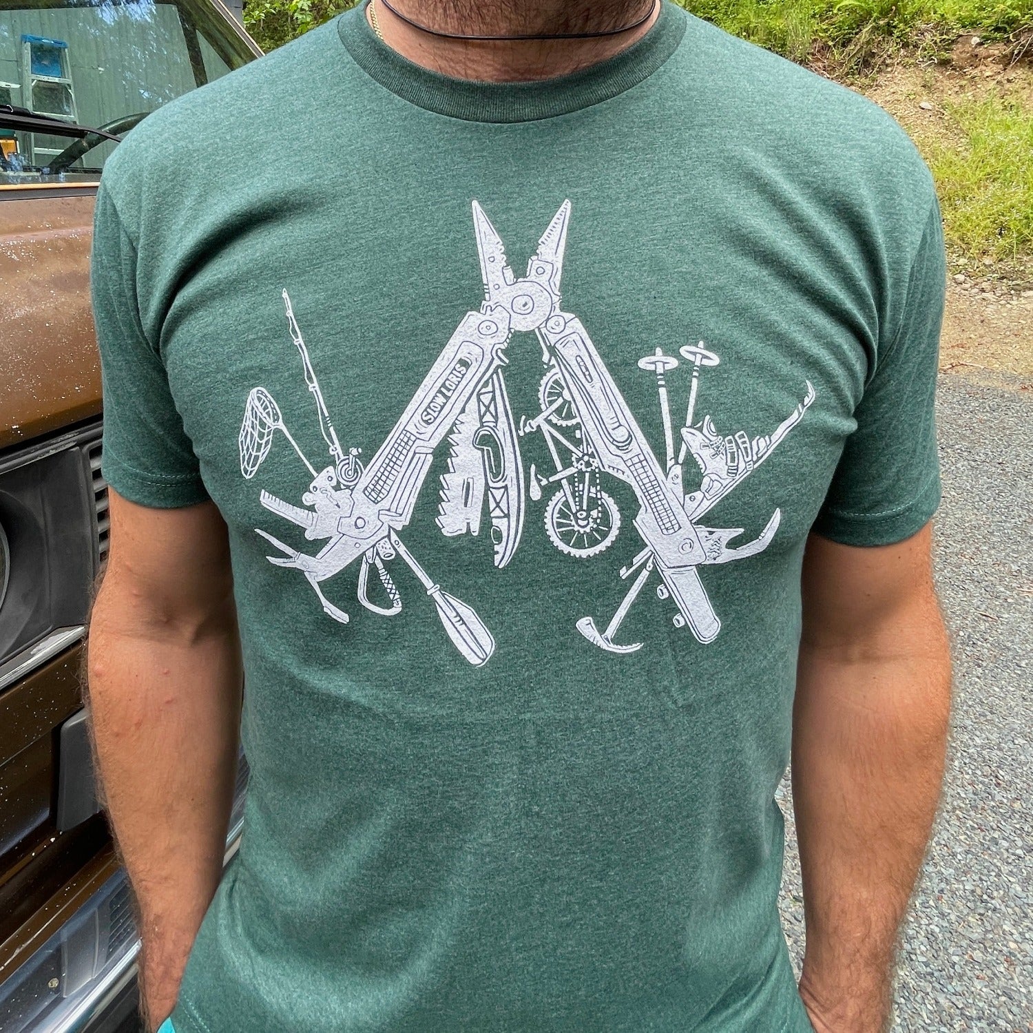 man wearing lighter green t-shirt with a white print of a multitool. The tools within the multitool are actually adventuring items- kayak, fishing pole, skis, ice axe, etc etc.