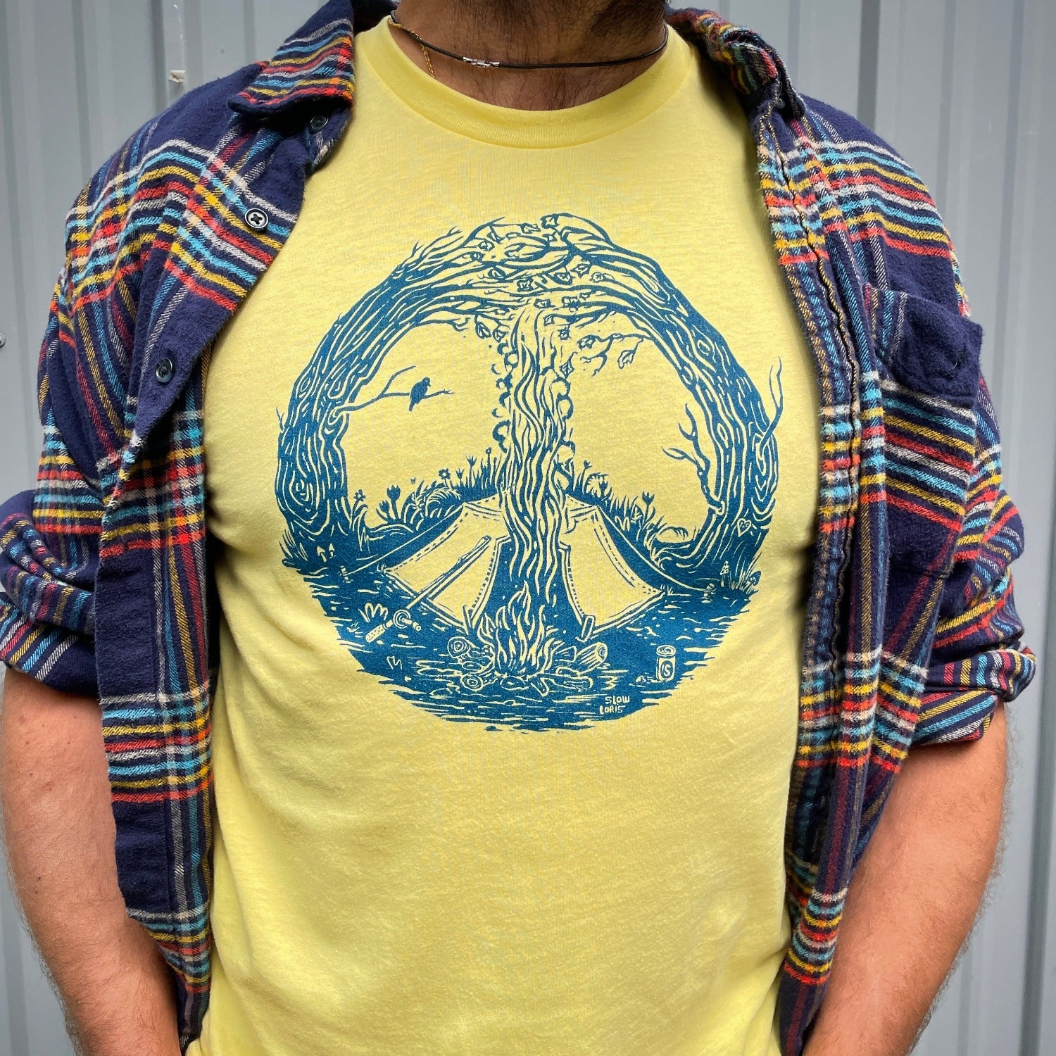 Close up of blue print on a yellow T. Print is of a campfire's smoke going up the center meeting with the tops of curved in trees creating a peace sign. Tent is by the fire and a bird is on a branch