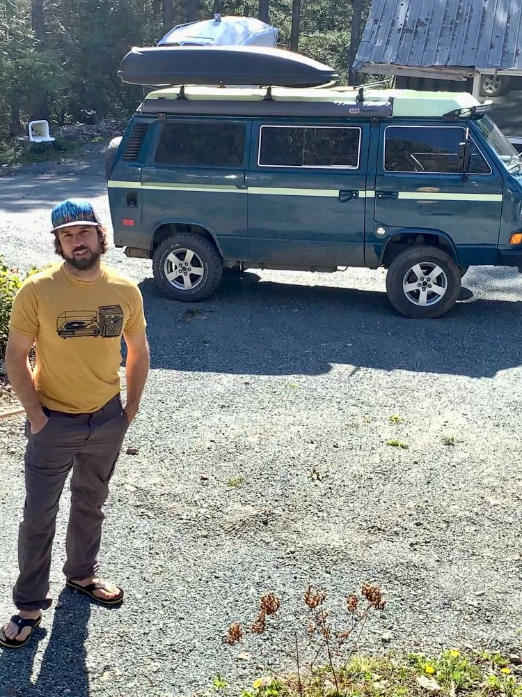 man standing by westfalia van wearing yellow shirt with black print of a record player with a milk crate full of records next to it.
