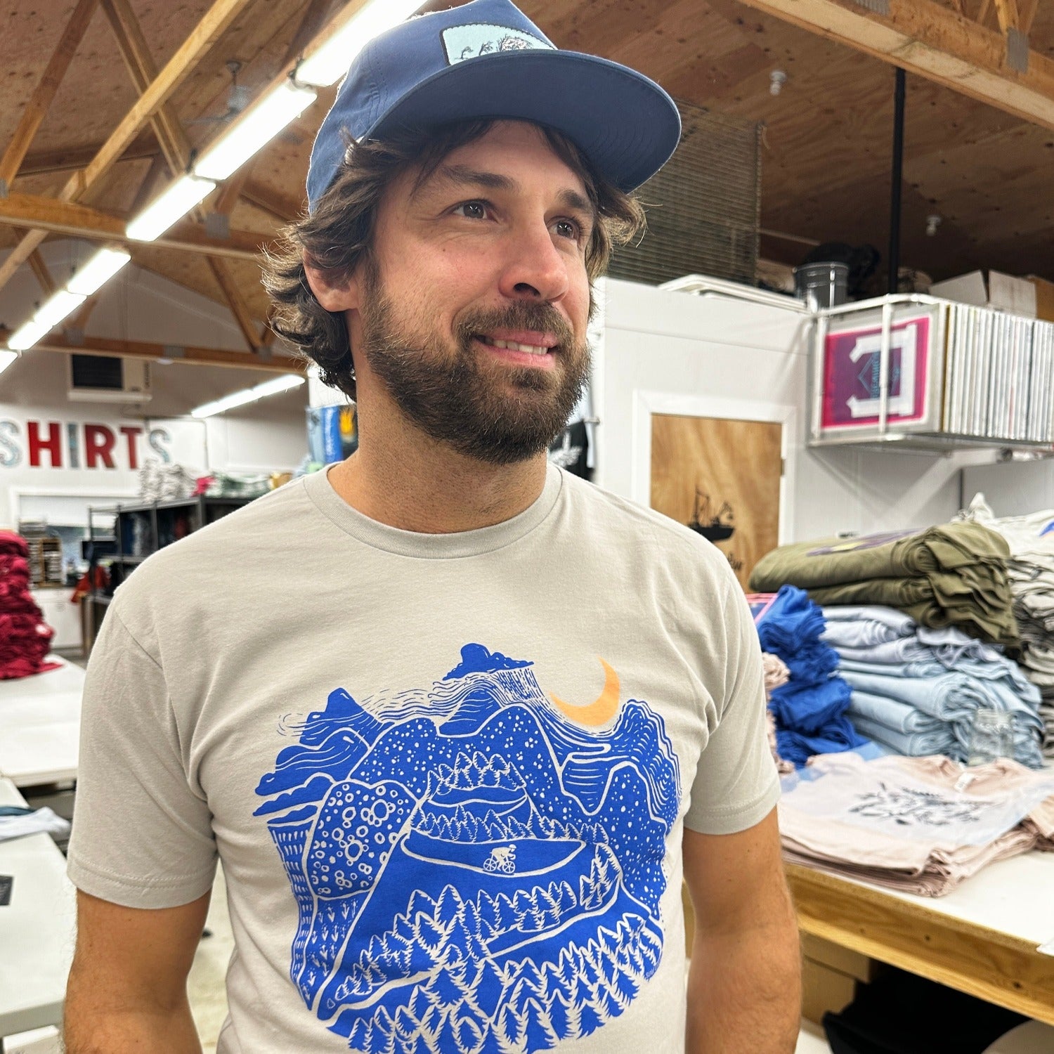Night Ride print on tan shirt in dark blue ink. Mountain scene with raincloud, crescent moon, and bicyclist riding down the mountain