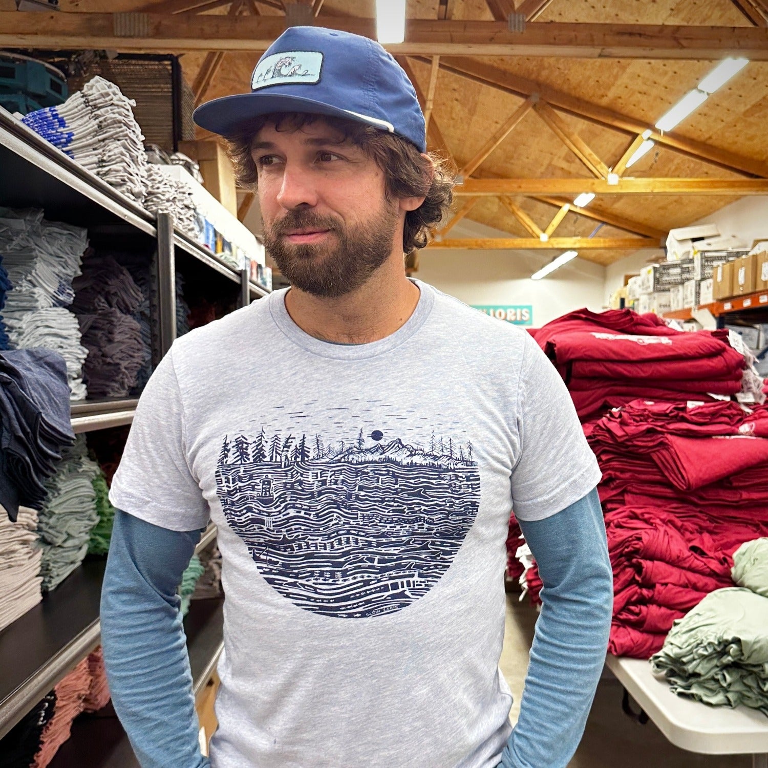 Man wearing heather light shirt with black ink print of the salish sea with mountains, trees, sky in the background.