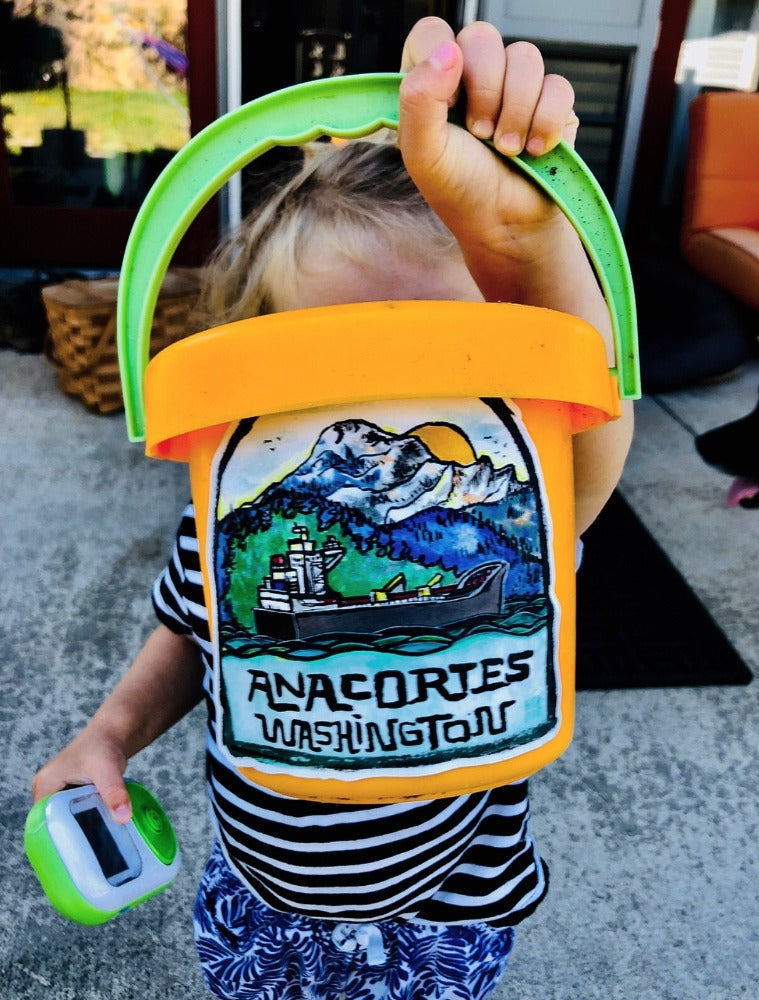 Colorfully painted sticker featuring marine landscape with words Anacortes, Washington underneath on child's toy beach bucket.