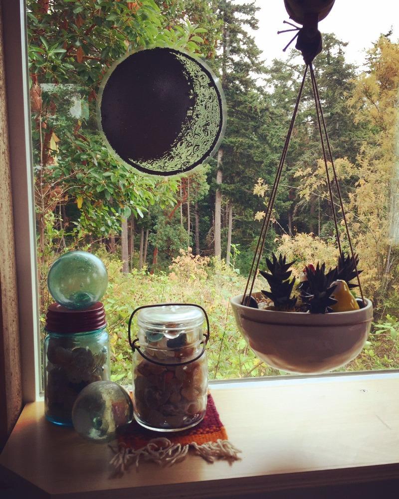 A large vinyl "stained glass" sticker in the shape of the waxing crescent moon, affixed to a window. Under the window are various jars and a hanging plant.