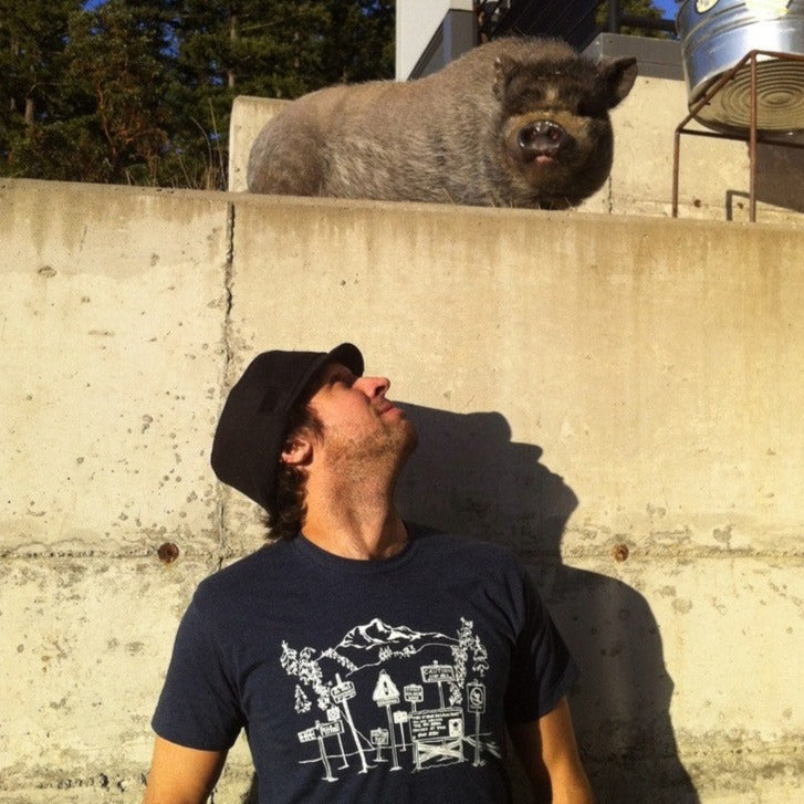 Man standing outside wearing blue t-shirt with white print of backcountry signs. A pig stands nearby.