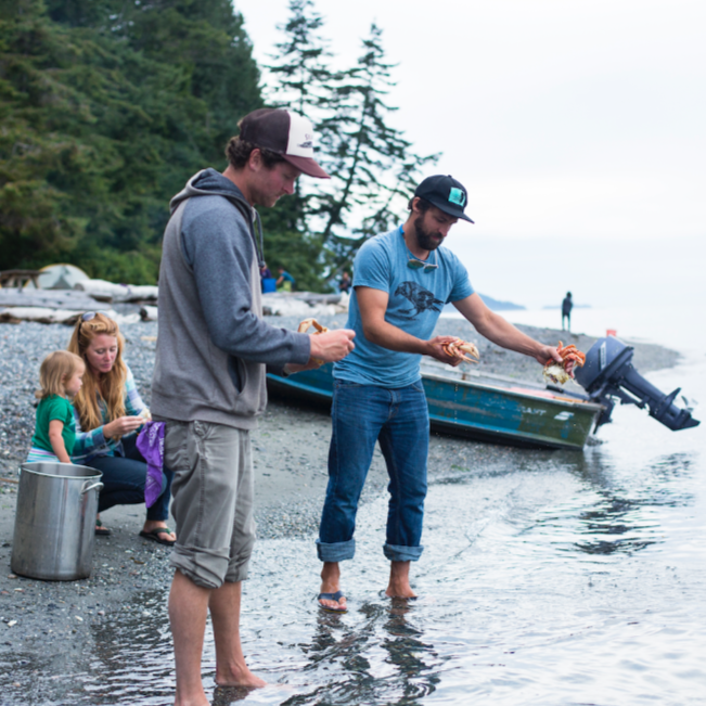 Blue t shirt screen printed with a black crow, worn by a man standing on the beach with his friends and family.