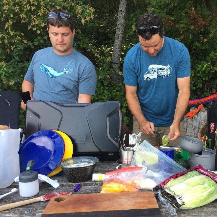 Two men cooking outside. One wears a t shirt printed with a blue whale, the other wears a t shirt printed with an old truck.