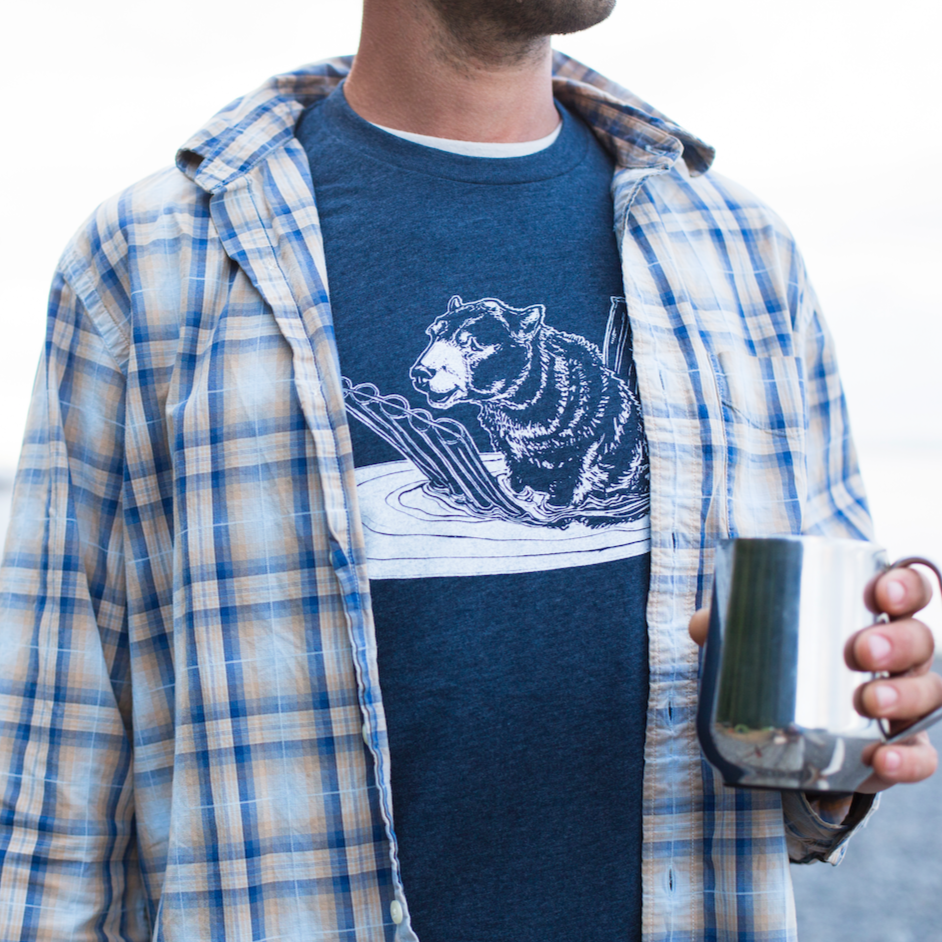 Man in flannel, holding a cup, and blue t shirt printed with a bear on a pool floatie.