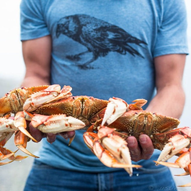 Blue t shirt screen printed with a black crow, worn by a man holding several cooked crabs.