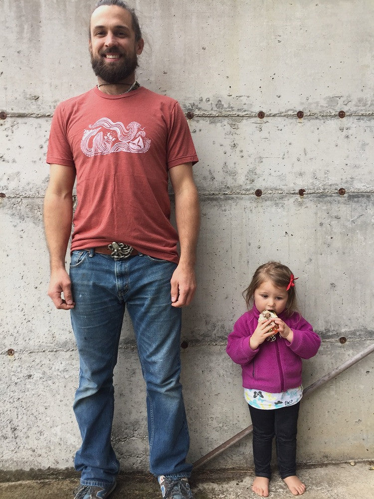 Man wearing clay t-shirt with white print of big winds and big seas tossing boats around. Next to model stands little toddler eating a huge sandwich