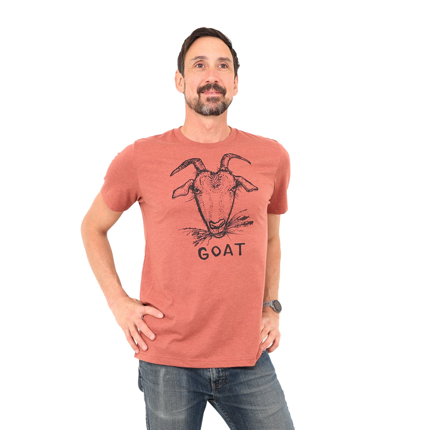 Man standing in white background, hands on his hips, wearing a clay colored shirt with a goat on it along with the word "goat"