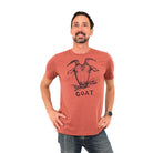 Man standing in white background, hands on his hips, wearing a clay colored shirt with a goat on it along with the word "goat"