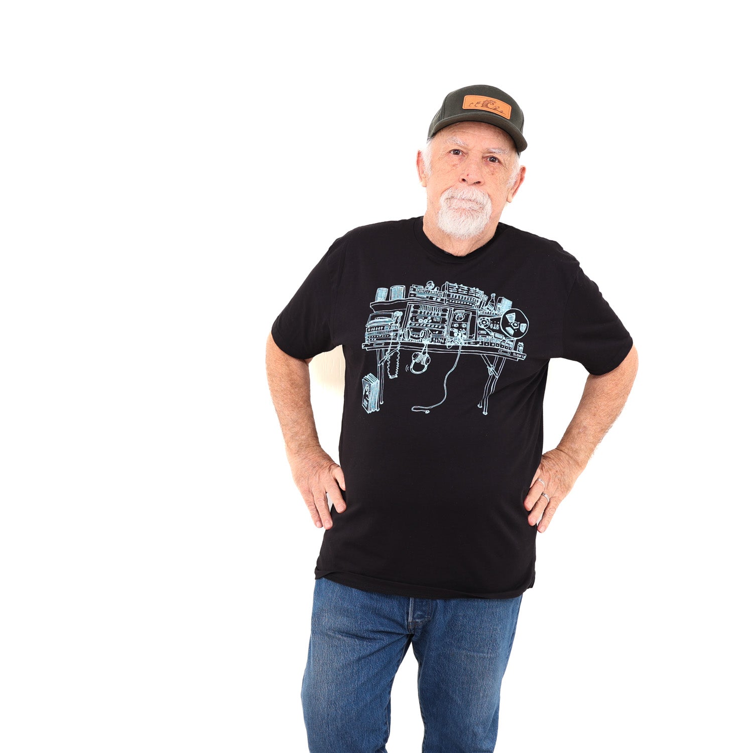 Man wearing hat standing with hands on his hips in a white background. Man is wearing black t-shirt with light blue ink of a recording studio