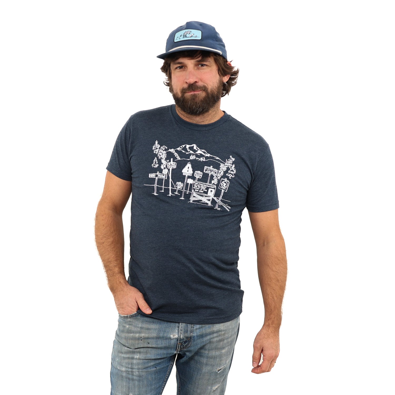 Man with dark hair and a beard wearing a blue shirt with danger signs saying to not ski that way with ski tracks going that way. Also wearing tattered blue jeans and a ball cap with trees turning to waves on it