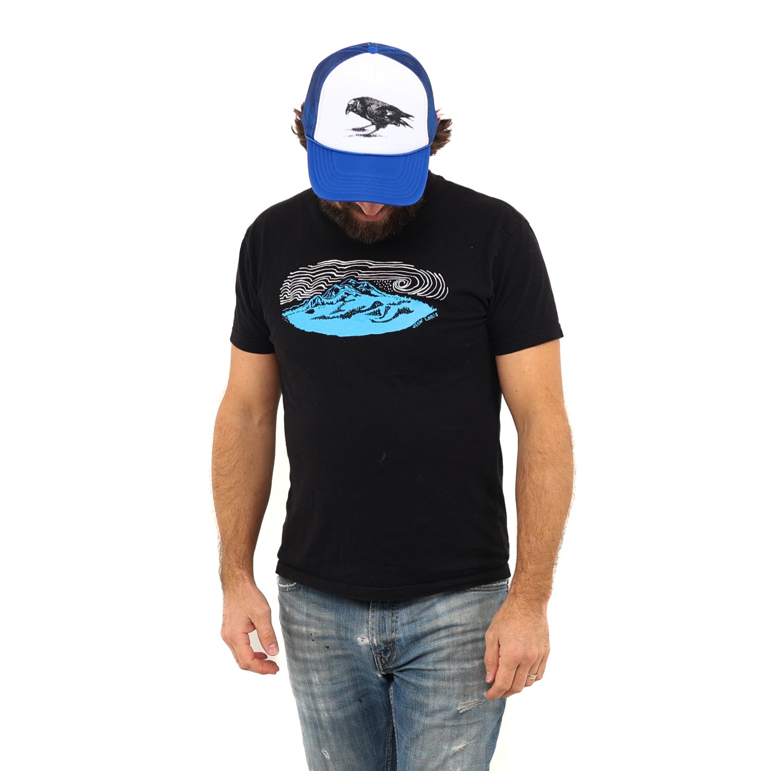 Man looking down wearing a ball cap in blue jeans and a black t-shirt with a blue mountain and white wind lines screenprinted 