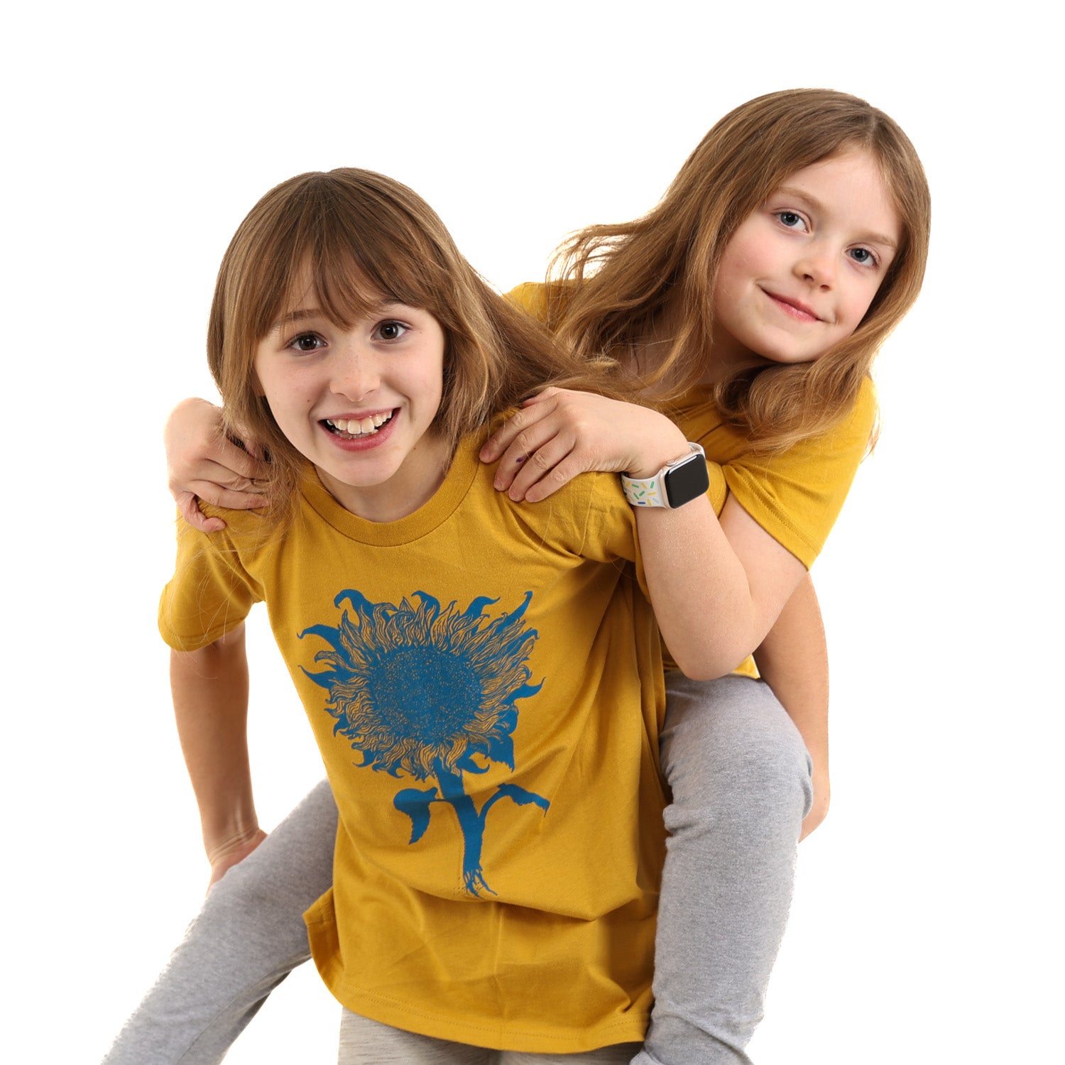 two little girls, one giving the other a piggy back, both wearing a mustard colored t-shirt with light blue print of an intricate sunflower.