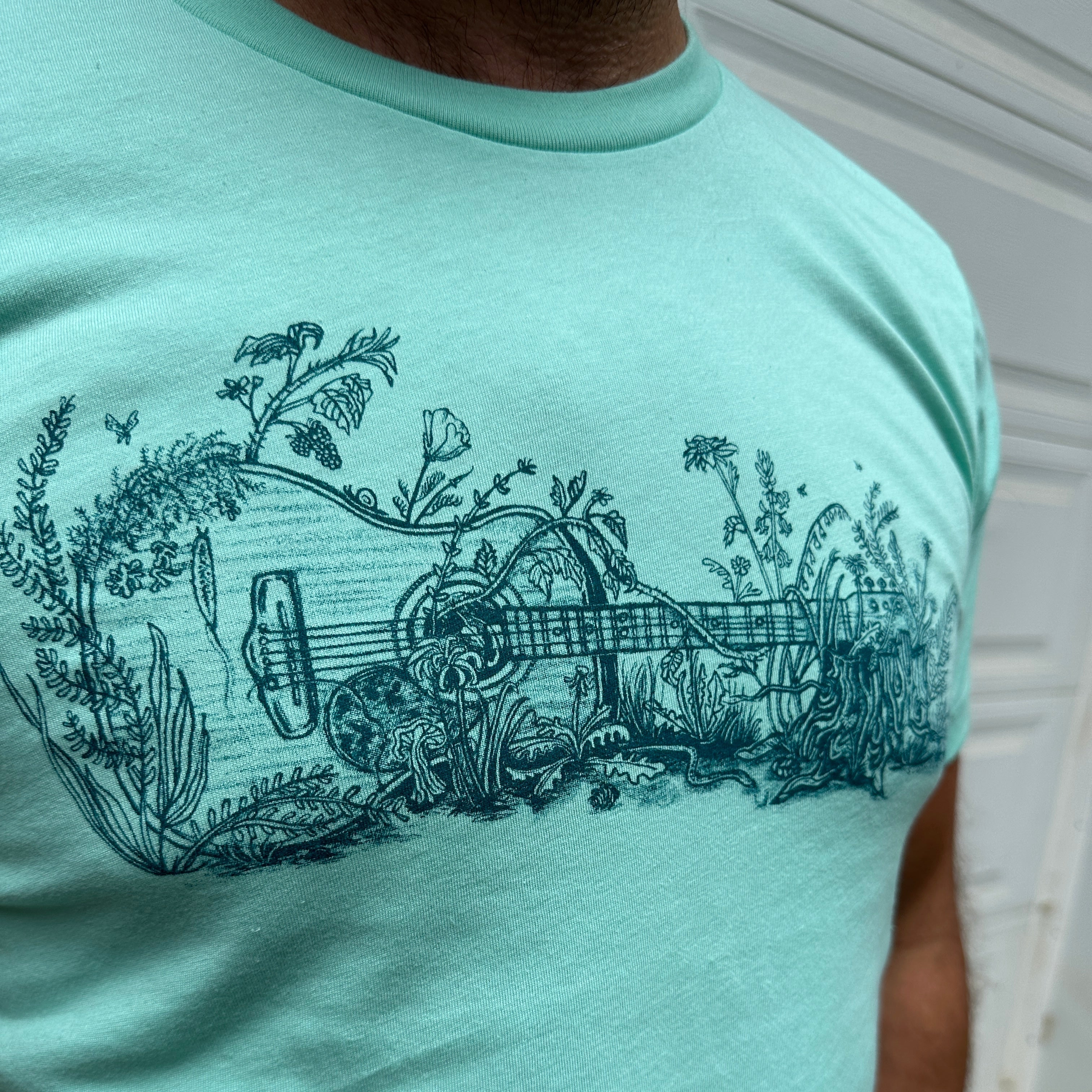 A drawing of a guitar on its side, overgrown with plants, printed in black ink on a light green t shirt.