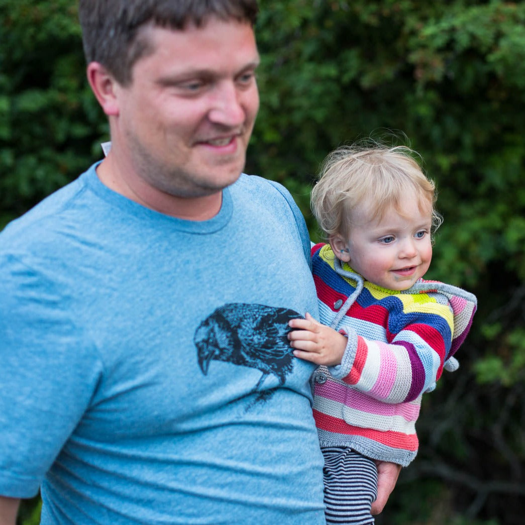 Man carrying his baby while wearing a blue shirt with a black crow on it. 