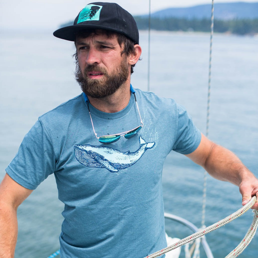 Man coiling a sailboat line wearing a blue shirt with a blue whale screen printed on it. Also wearing a ball cap and sunglasses around neck 