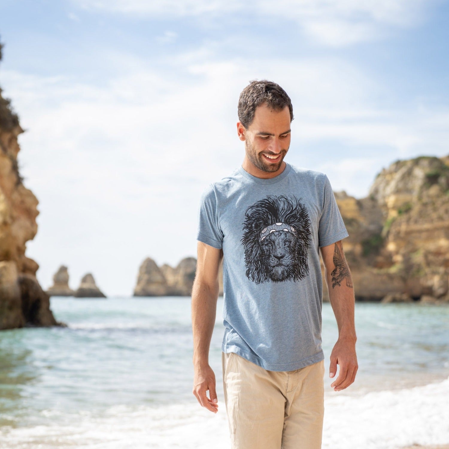 Man on beach with water and rocky coast in background. Man is wearing a steal blue tee with a screen printed lion wearing a headband on it. 