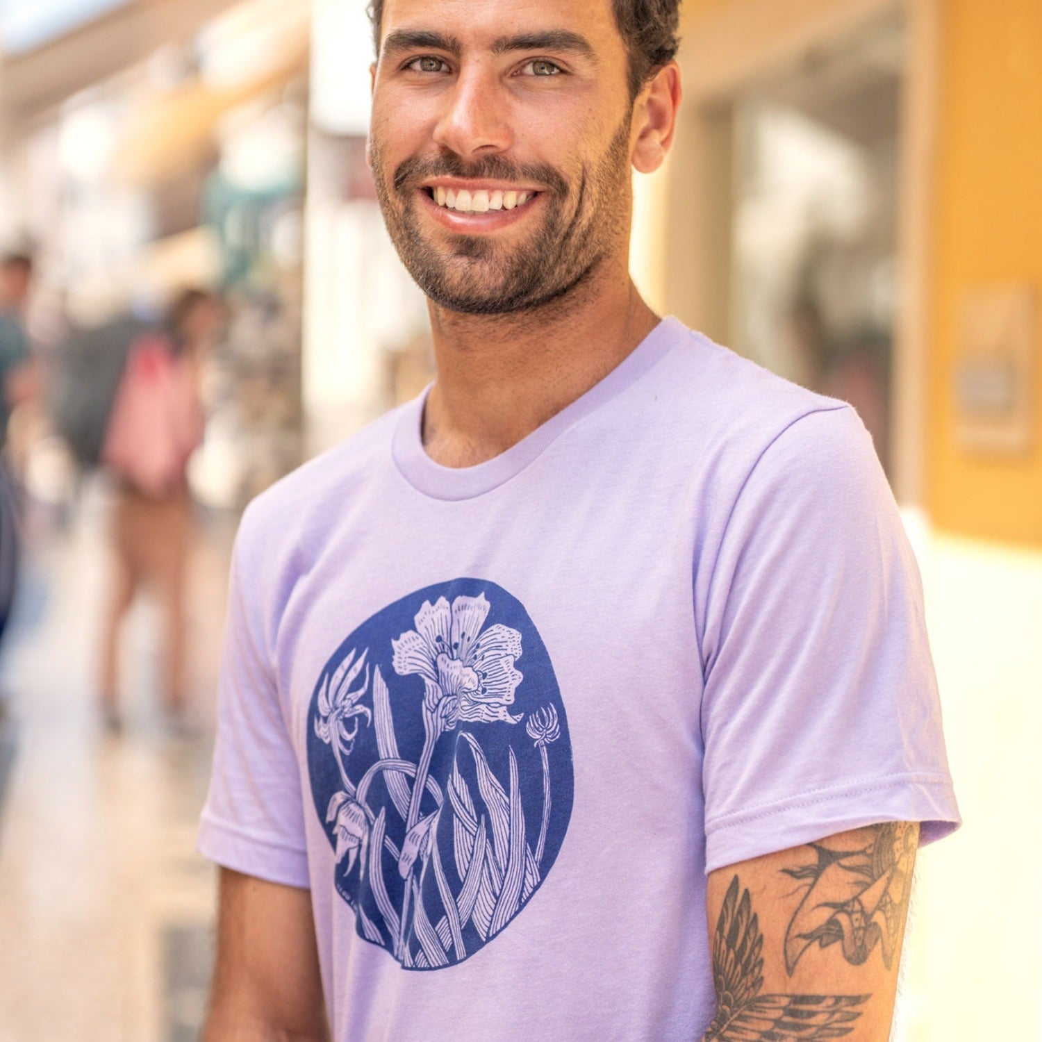 Man smiling wearing a light purple shirt with dark purple flower print on it. People in background out of focus.
