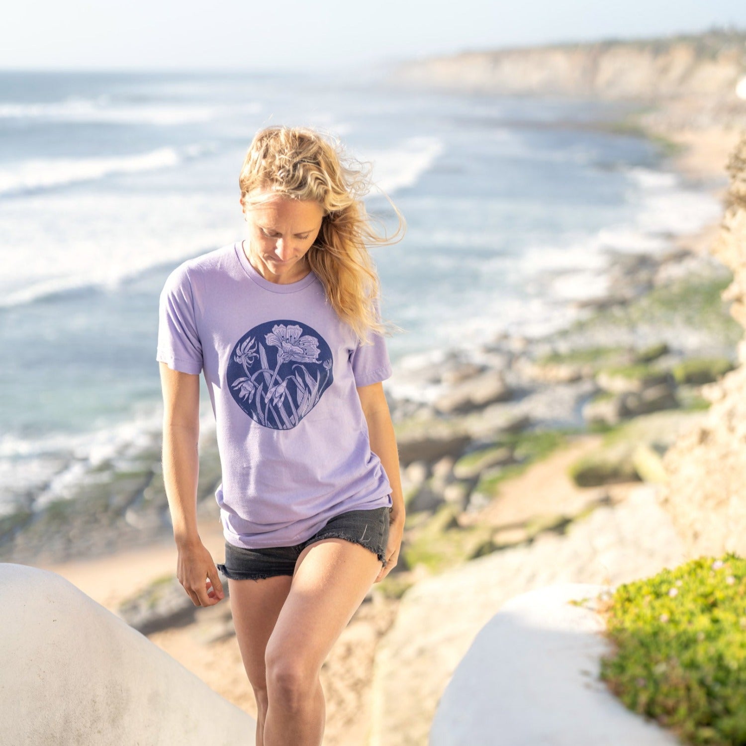 Girl walking up stairs with a beautiful beach scene in the distance. Girl has blond hair and is wearing a light purple shirt with a dark purple flower print on it. 