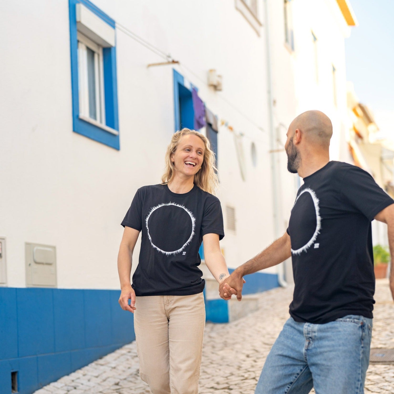woman and man walking down cobblestone street with blue building behind. Couple is holding hands and smiling at each other wearing the same black shirt with an eclipse print in white. 