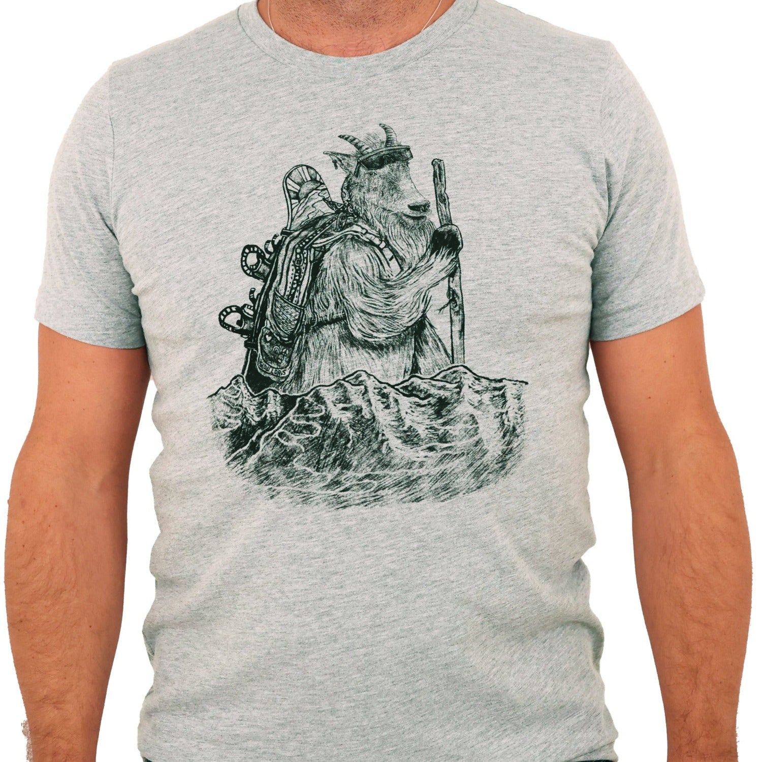 heather grey t-shirt with black ink of a mountain goat hiking with a walking stick, sunglasses, and a snowboard on his backpack. Mountains with fresh tracks anterior