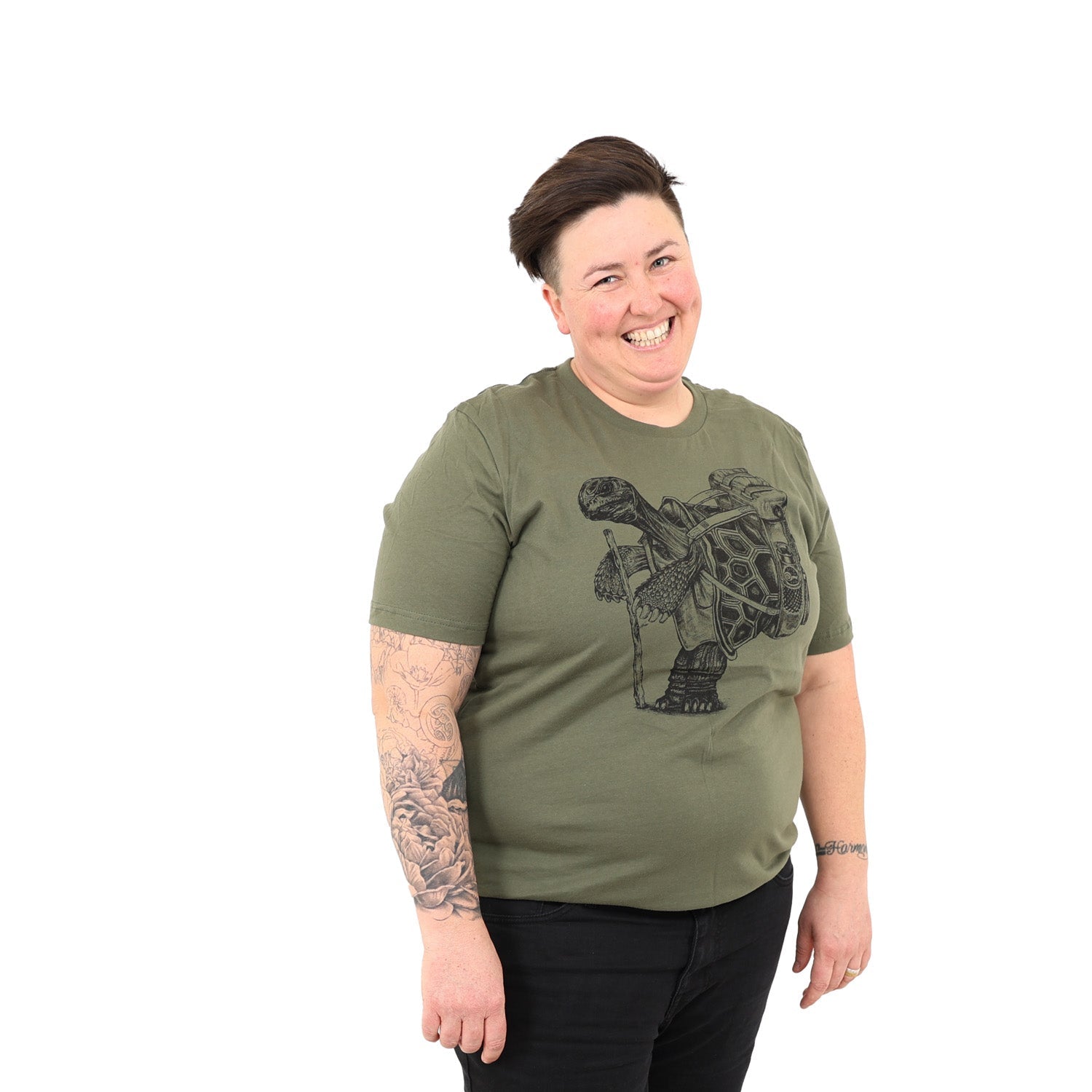 woman with short hair smiling with a white background while wearing an army green shirt with a screen printed tortoise standing upright wearing a back pack holding a walking stick