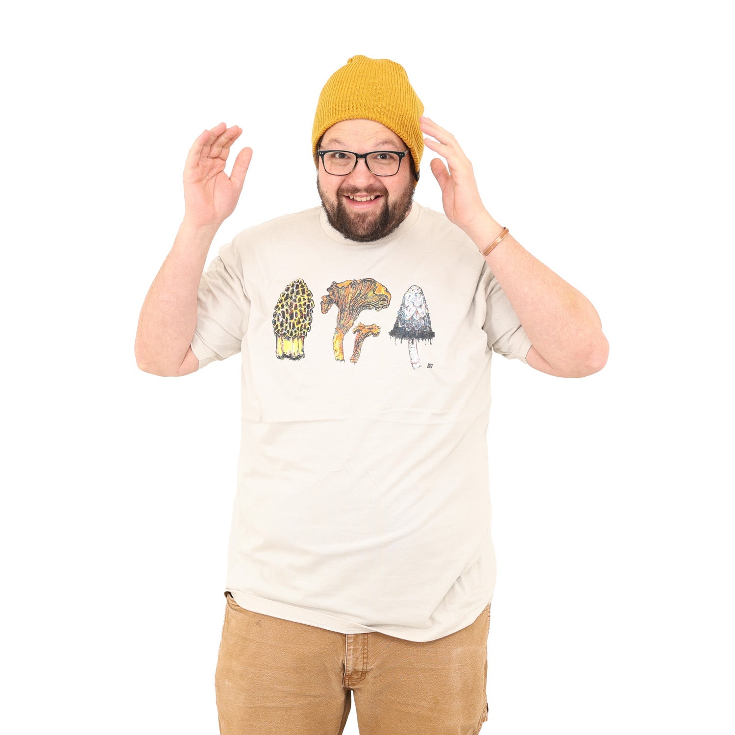 Man with his hands up by his head, smiling, wearing glasses and a beanie. Man is wearing a tan colored shirt with three mushrooms- morel, chantrelle, and shaggy mane