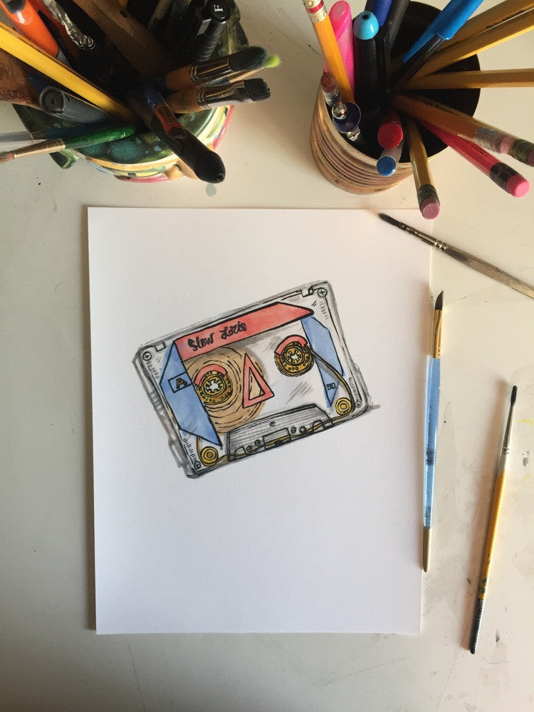 Art print of colorful cassette tape against white background, laid on desk with paintbrushes and pencils