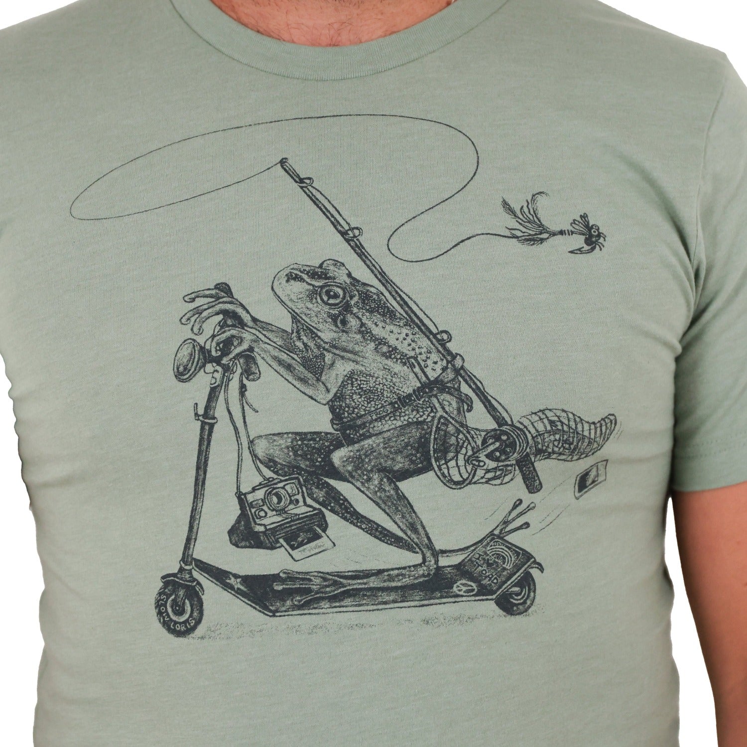 man torso wearing green shirt with print of a frog riding a scooter with a fly fishing rod, net, polaroid camera