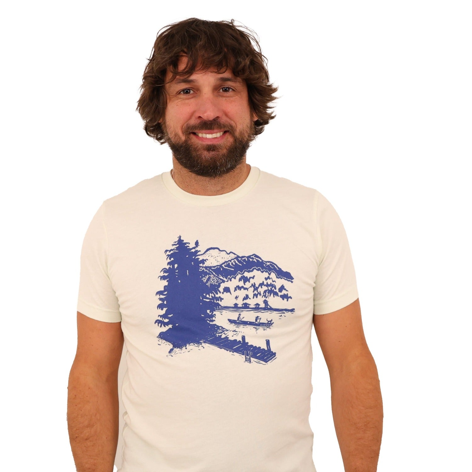 Man wearing citron(light tan) t-shirt with blue print of lake scene with a dock, canoe, trees, mountains, etc.