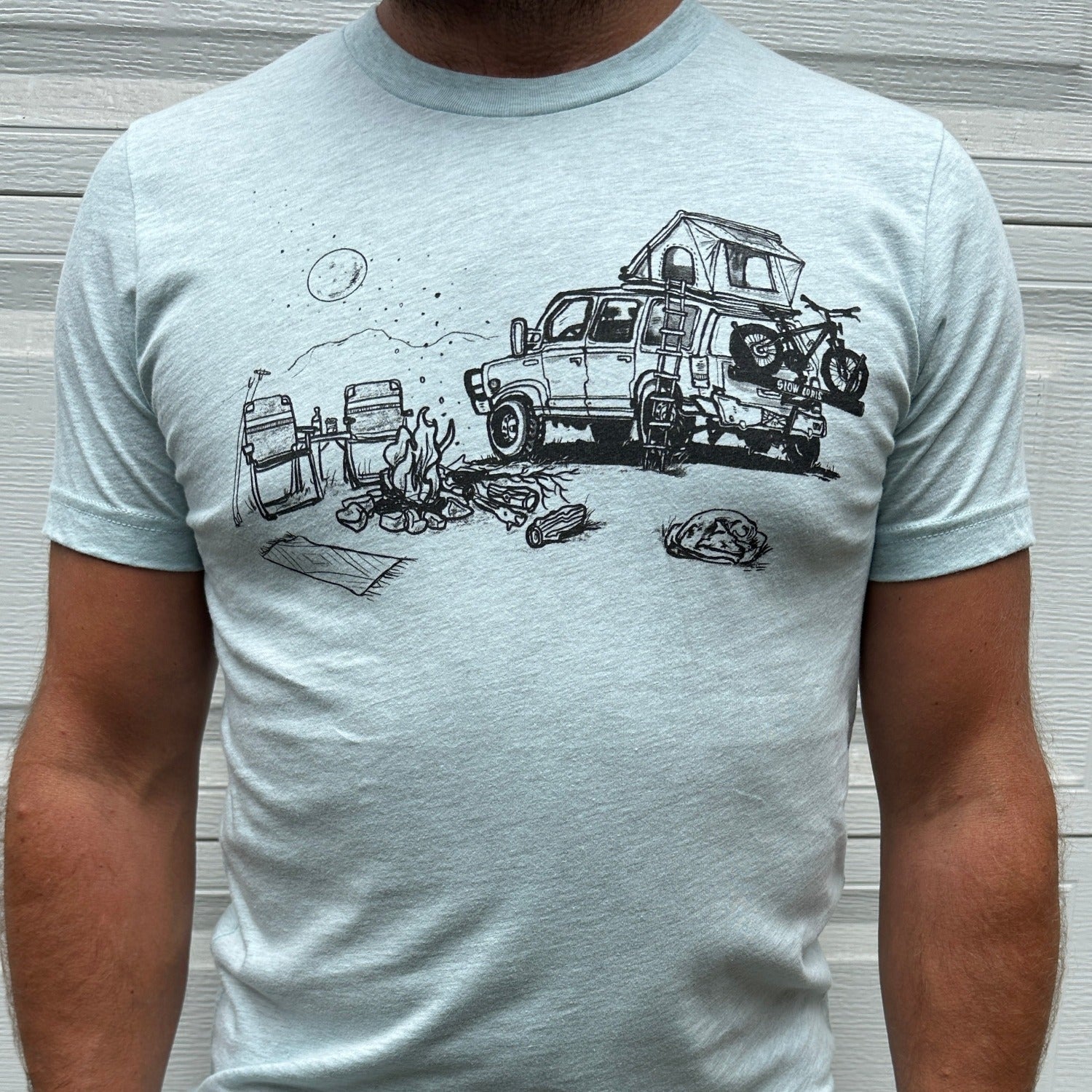 Man wearing light blue shirt with black ink print of campfire scene; car with rooftop tent with ladder, bike on the back. Campfire, two chairs, mountains and full moon. 