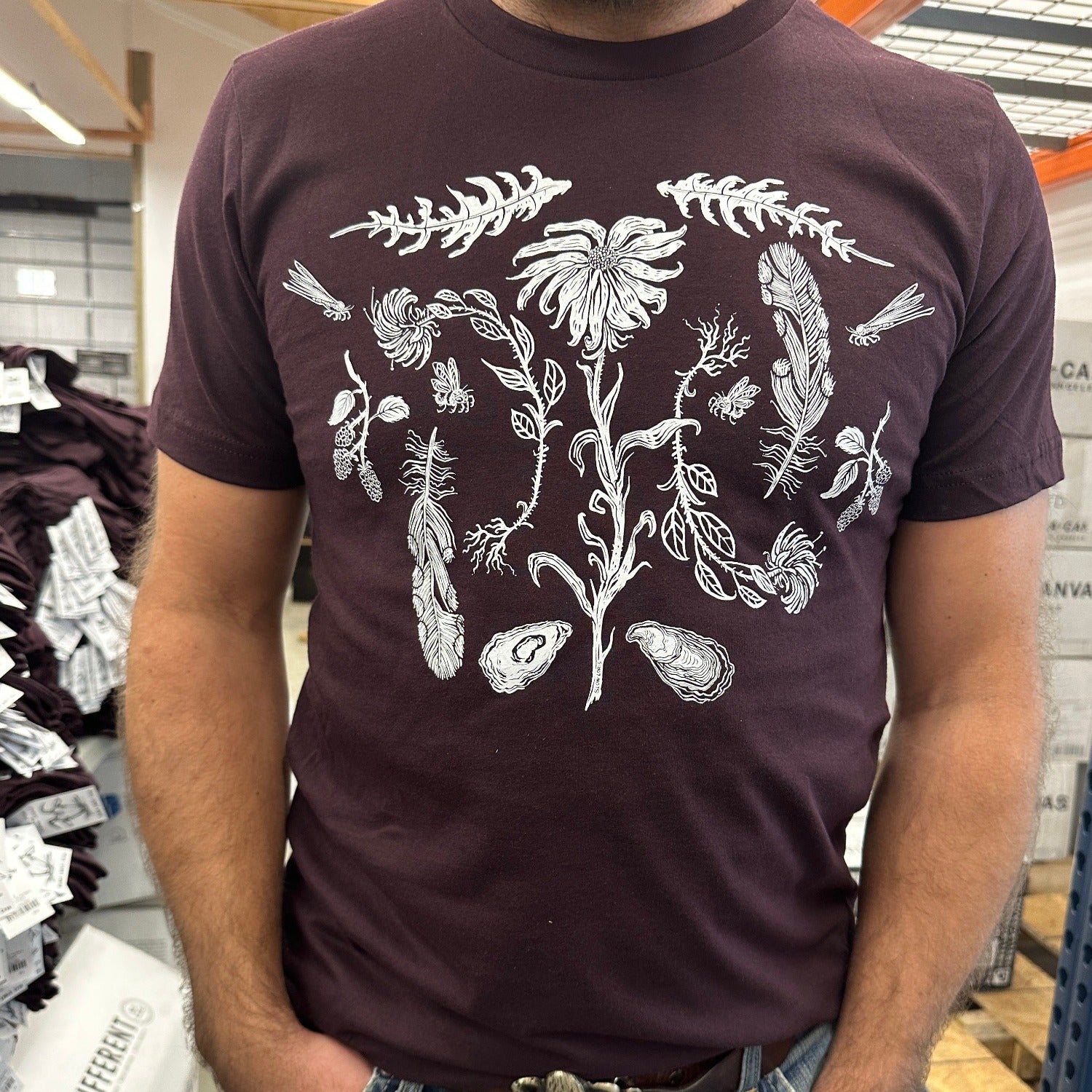 close up of a oxblood colored t-shirt with white ink of flowers, ferns, feathers, shells, etc