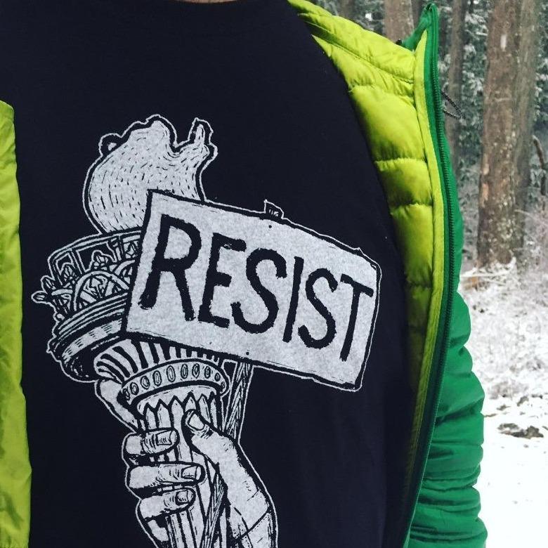 man outside with snowy background wearing black resist t shirt and jeans with a green puffy zip up