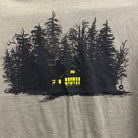 close up showing print of brown t-shirt with print of black ink of silhouetted trees with yellow ink house lights shining out of darkness. Two animals sitting in the window.