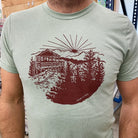 Close up of a soft green t-shirt with maroon red print of a lookout tower in the mountains. 