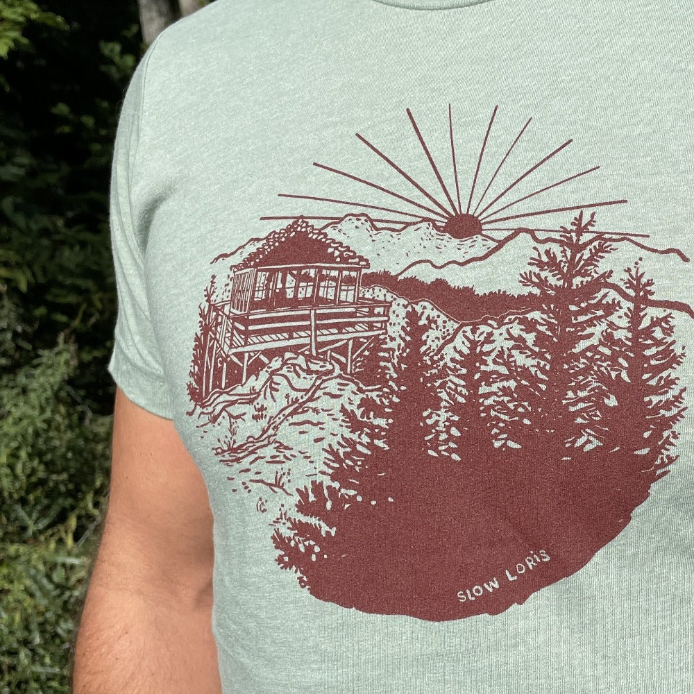 Close up of a soft green t-shirt with maroon red print of a lookout tower in the mountains.