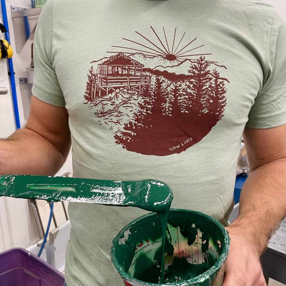 Close up of a soft green t-shirt with maroon red print of a lookout tower in the mountains. Man is holding green ink in his hands. 