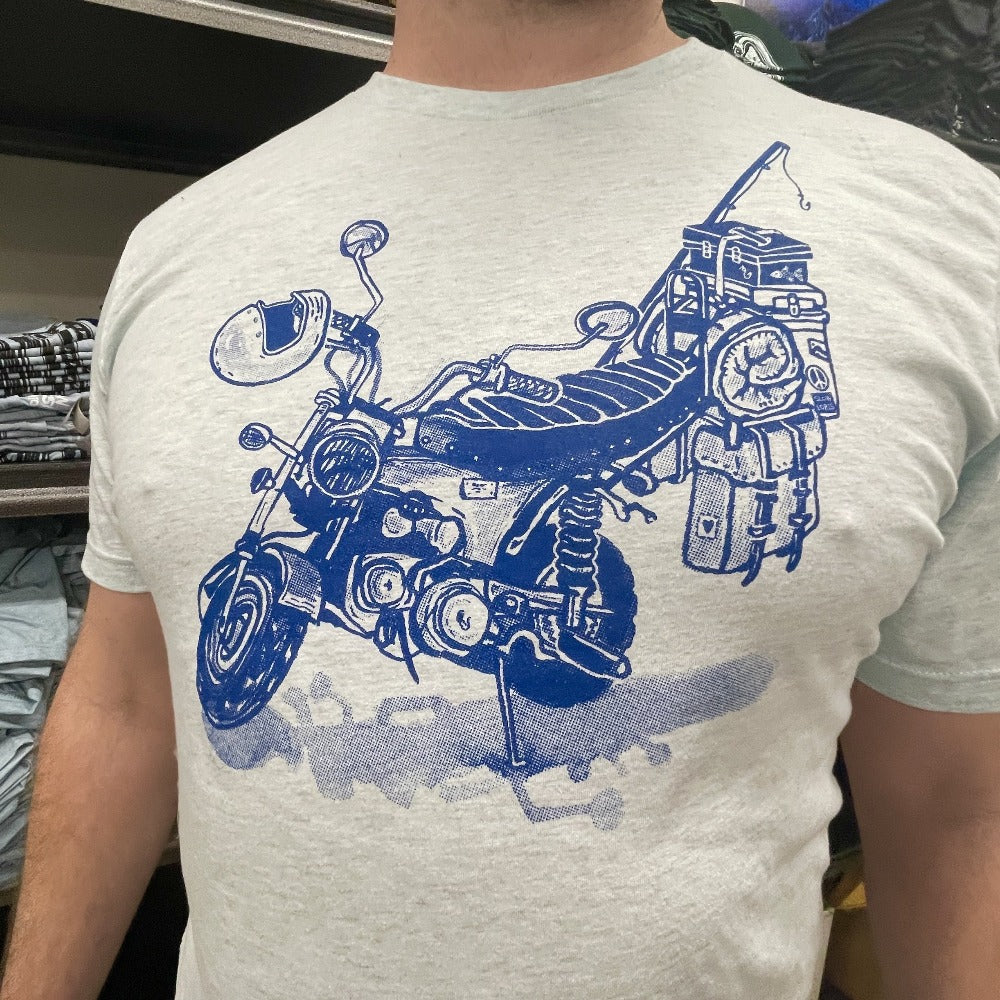 Blue print on a cream colored t-shirt. Print is of a mini-bike loaded up with fishing pole, helmet, sleeping bag, and bags.