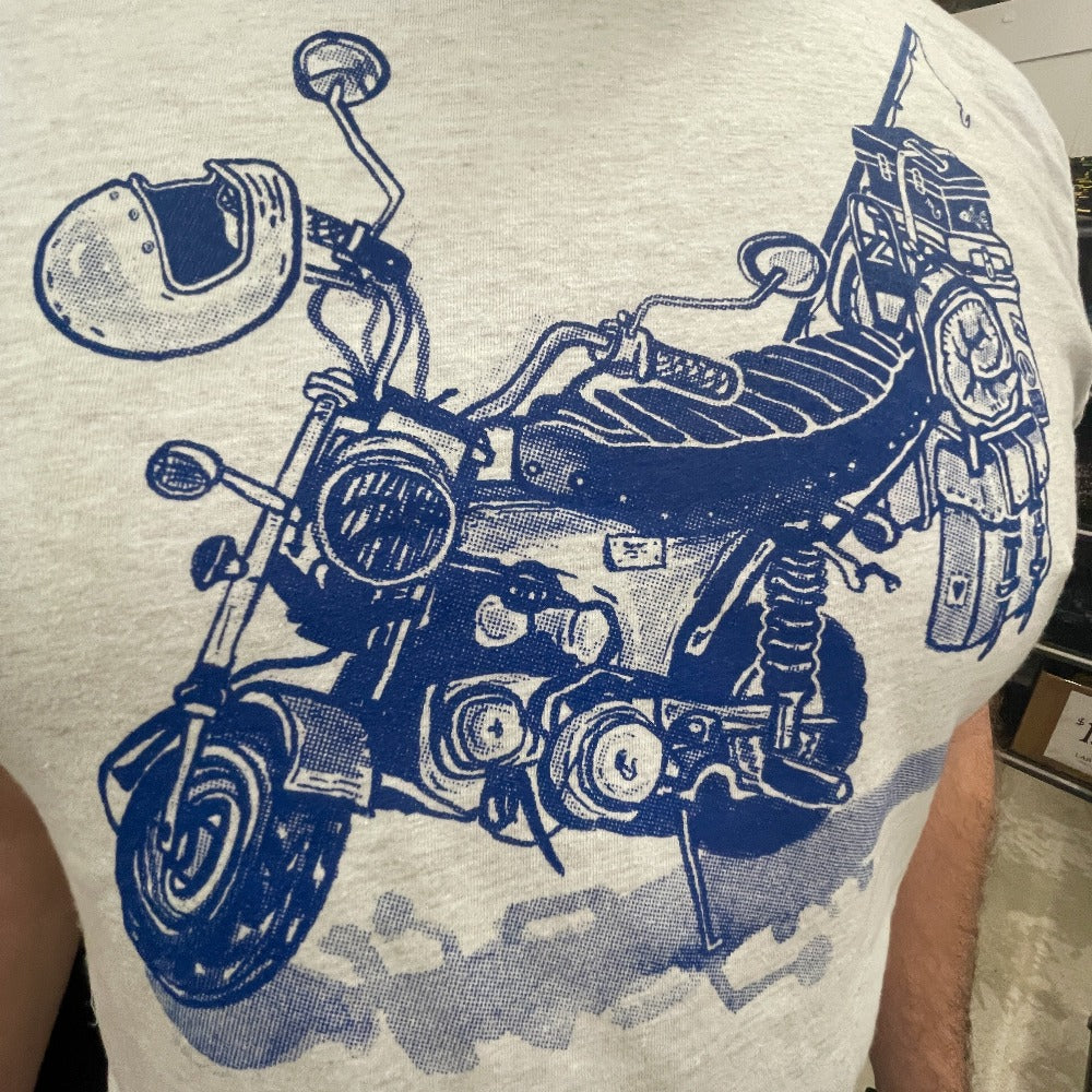 Blue print on a cream colored t-shirt. Print is of a mini-bike loaded up with fishing pole, helmet, sleeping bag, and bags.