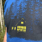 Close up of man wearing blue t-shirt with print of black ink of silhouetted trees with yellow ink house lights shining out of darkness. Two animals sitting in the window.