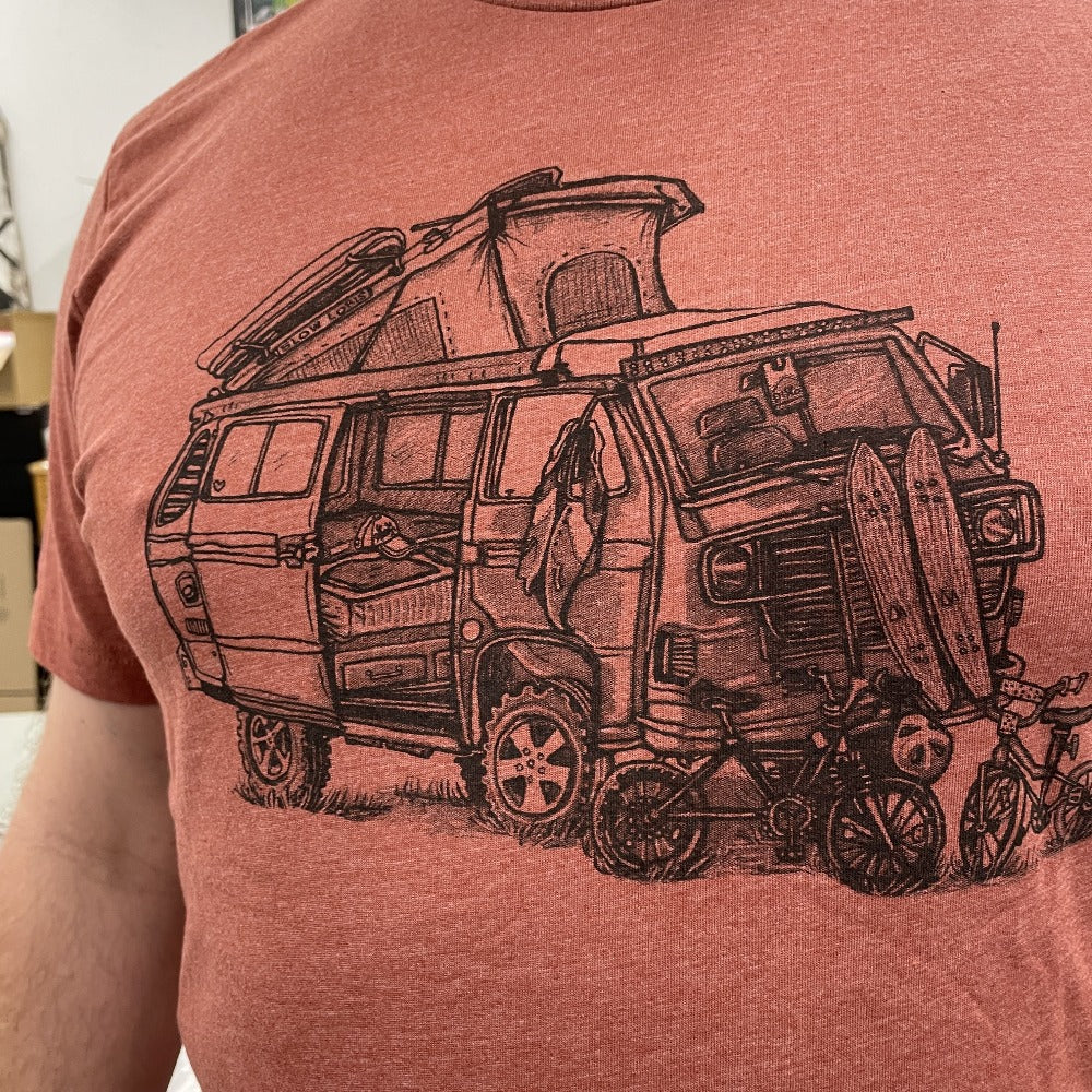 close up of man wearing clay colored shirt with a VW van open and top popped. Skateboards, bikes, and clothing strewn about. 
