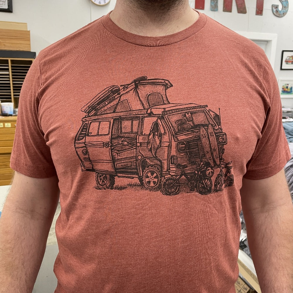 close up of man wearing clay shirt with a VW van open and top popped. Skateboards, bikes, and clothing strewn about. 