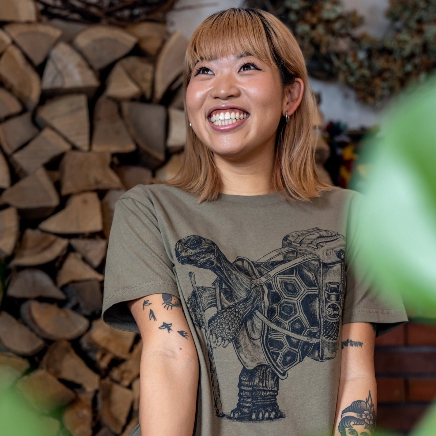 Woman smiling really big. Stacked split wood in the background. Tattoos on her arms. Wearing an army green t-shirt with dark blue ink of a hiking tortoise.  