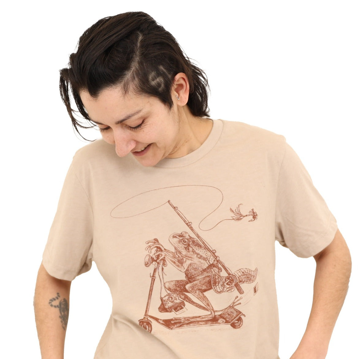 woman wearing tan shirt with print of a frog riding a scooter with a fly fishing rod, net, polaroid camera
