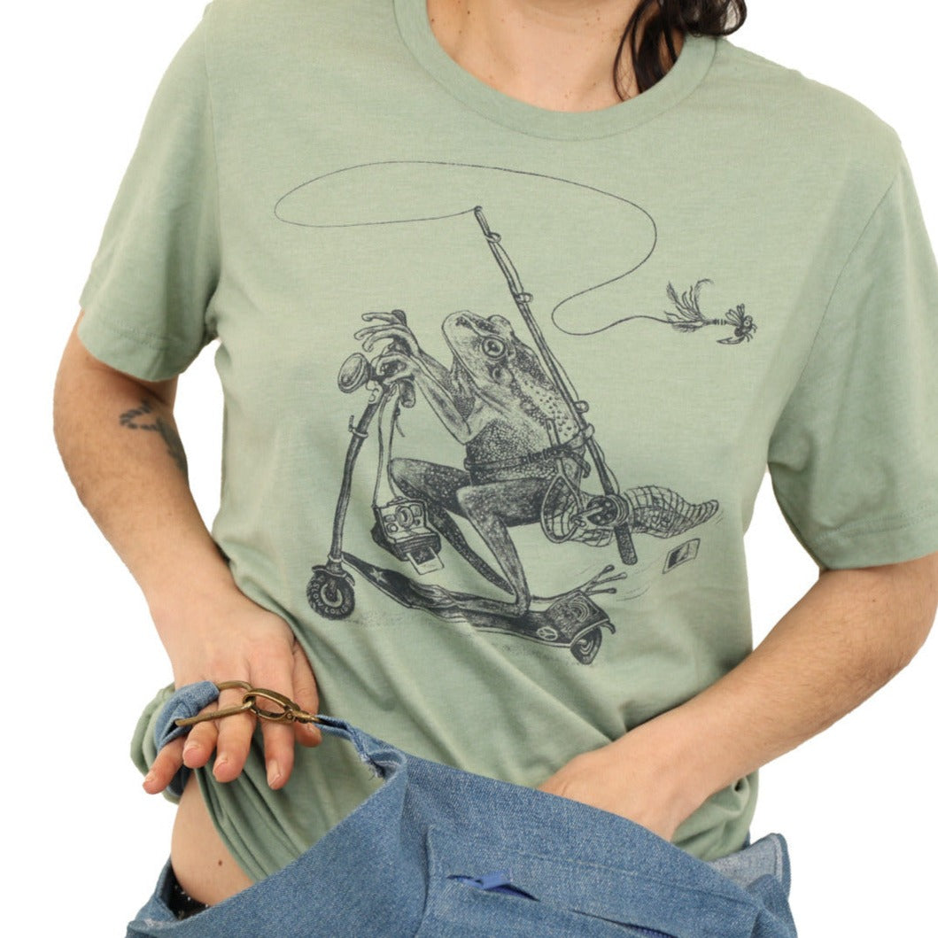 woman wearing green shirt with print of a frog riding a scooter with a fly fishing rod, net, polaroid camera