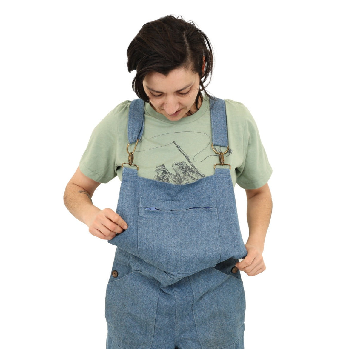 Woman in overalls wearing a green shirt with print of a frog riding a scooter with a fly fishing rod, net, polaroid camera
