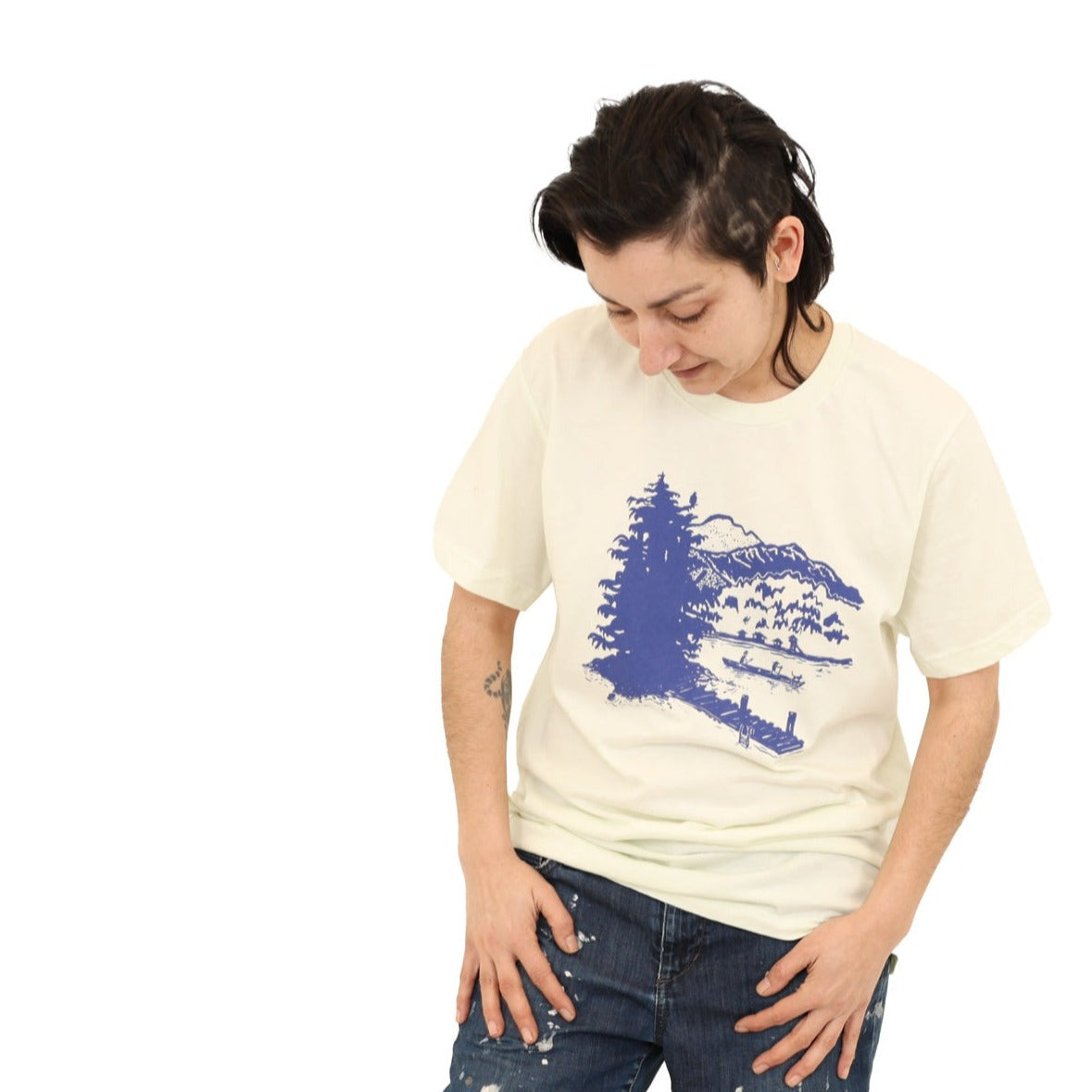 Woman wearing citron(light tan) t-shirt with blue print of lake scene with a dock, canoe, trees, mountains, etc.