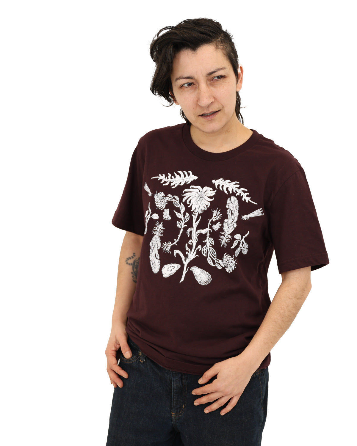 Woman wearing a oxblood colored t-shirt with white ink of flowers, ferns, feathers, shells, etc